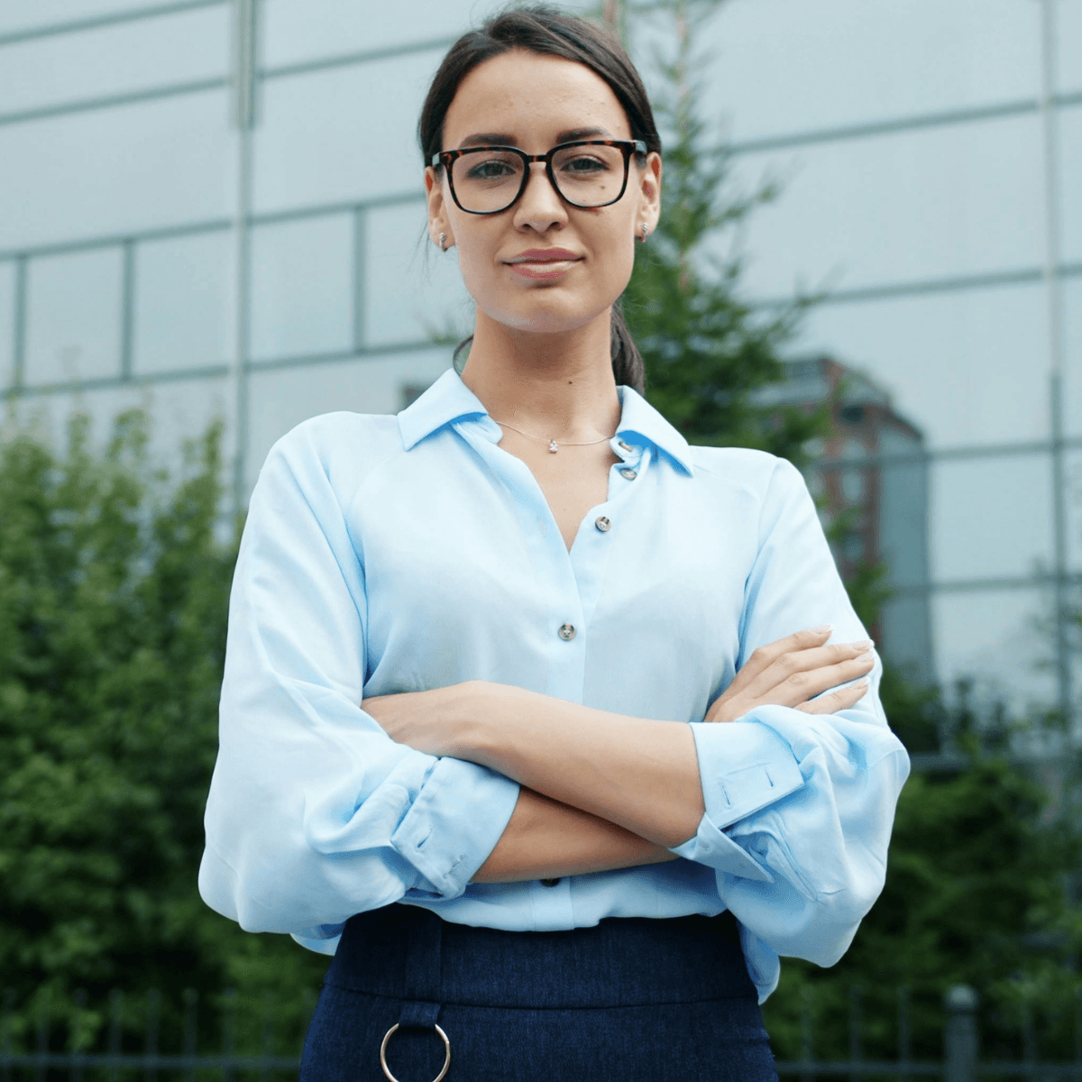 Professional woman wearing glasses and a light blue blouse stands with arms crossed outdoors.