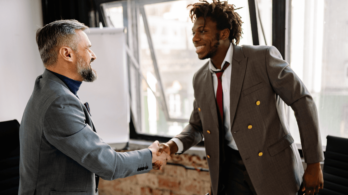 Smiling older white businessman and younger Black man with dreadlocks shaking hands in an office.
