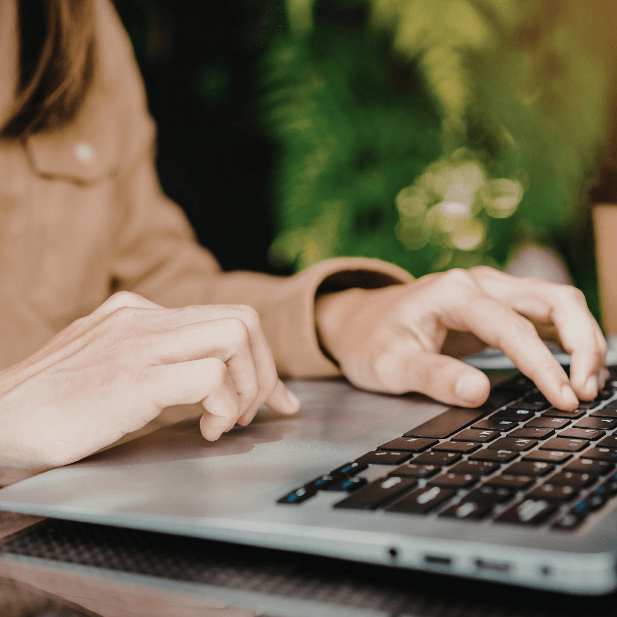 Close-up of hands typing on a laptop keyboard with blurred green foliage in the background.
