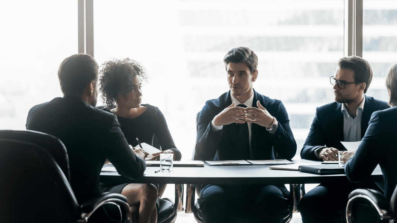 Diverse business professionals in suits sit at a conference table, listening to a colleague speak.