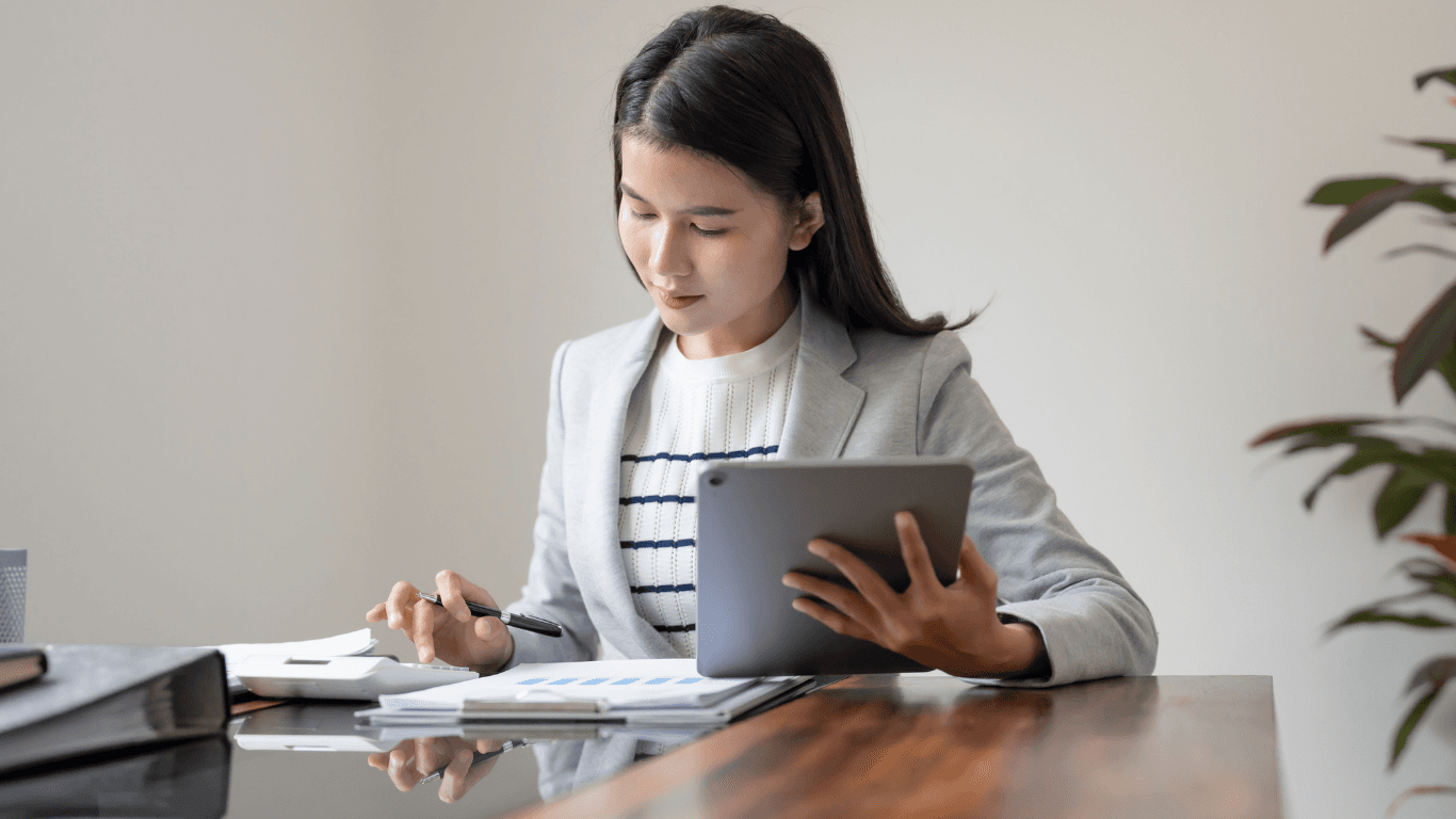 Focused businesswoman using a tablet and calculator to review financial documents at her desk.