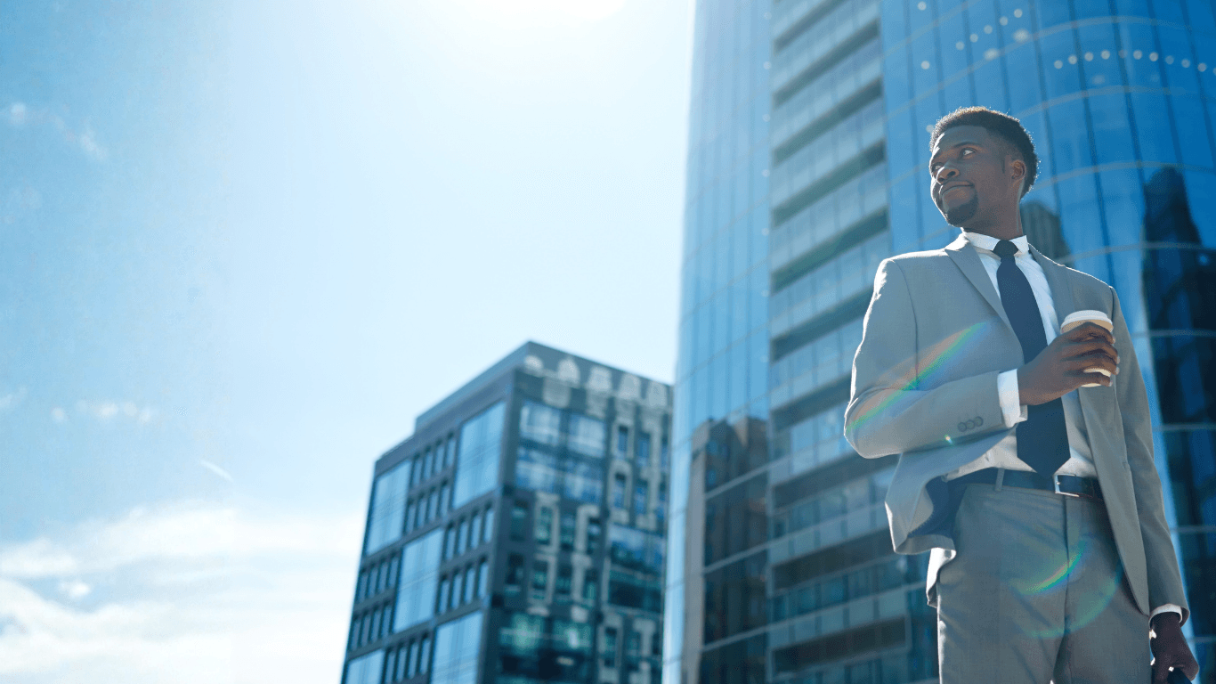 Black businessman in a grey suit holding coffee, standing before modern glass skyscrapers in sunlight.