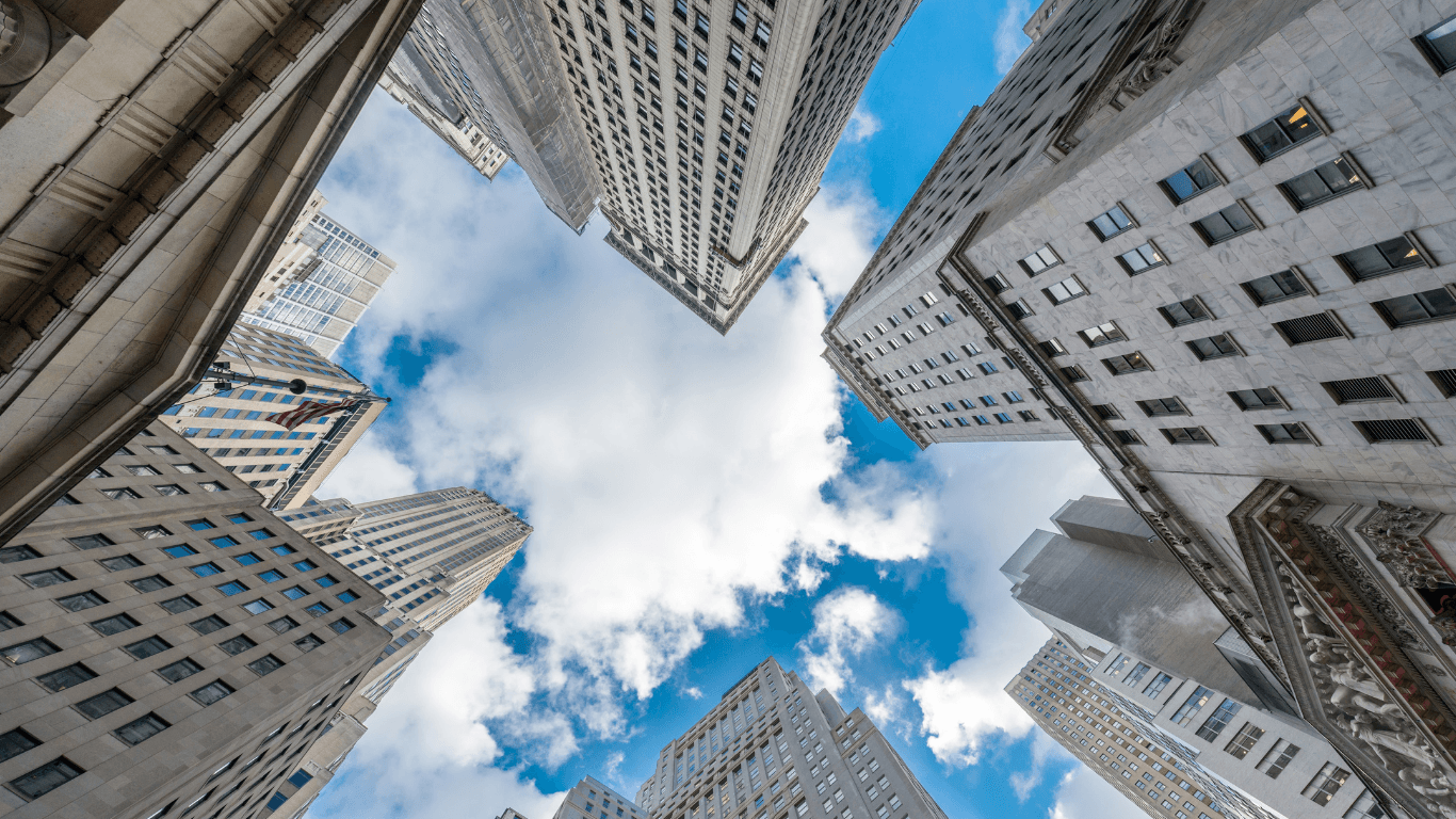 Towering urban skyscrapers reach toward a bright blue sky filled with soft white clouds.